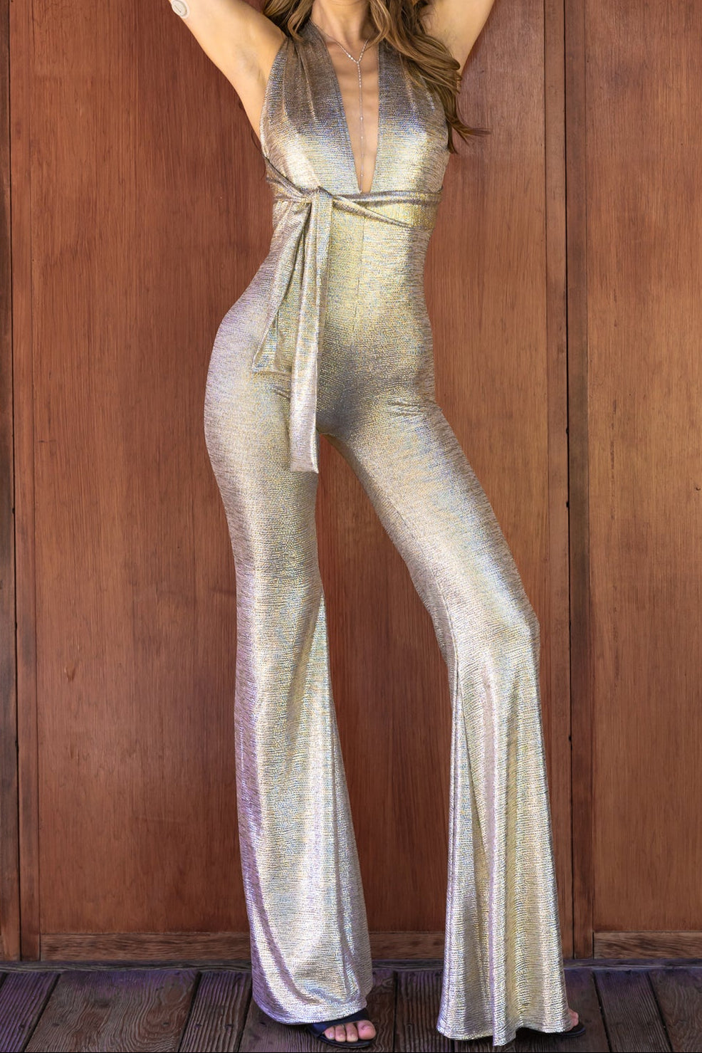 A woman modeling a shimmering metallic silver halter jumpsuit with a deep V-neckline and tie waist, posed against a wooden plank wall.