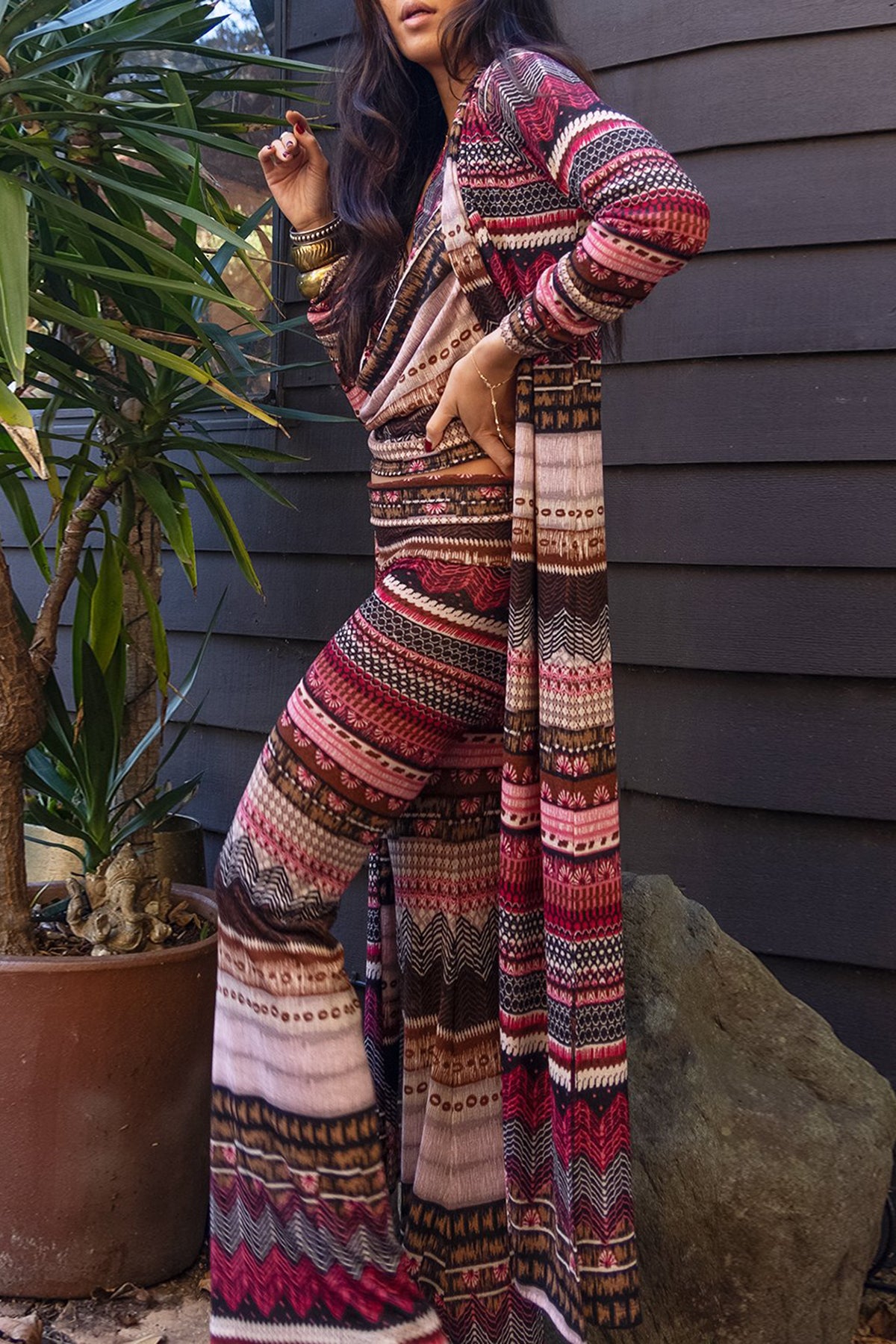 A person wearing a multicolored maxi-length lounge robe with a geometric pattern, featuring long sleeves and a side slit, standing near a house and a potted plant.