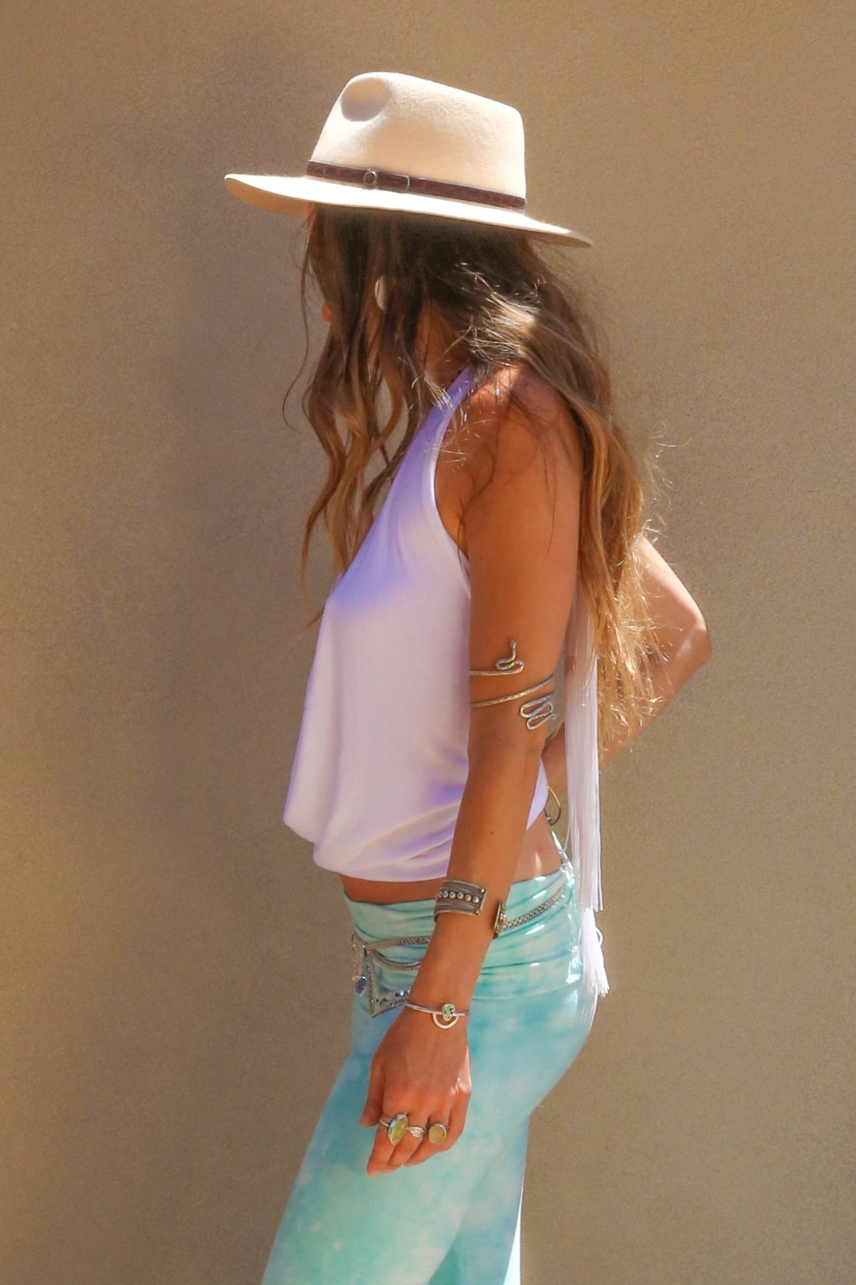 Woman wearing a white sleeveless top and light blue pants with a beige hat against a beige wall.