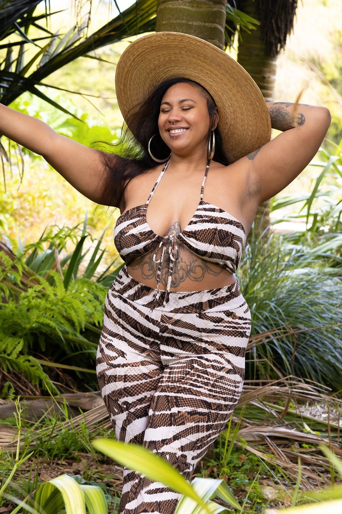 Model in a brown and tan animal print outfit and wide brimmed hat standing in a jungle setting